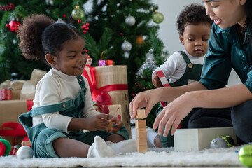 Happy mixed race family, little African kids girl and boy with green bib and Asian young mother playing wooden blocks together at home on Christmas holiday. Xmas tree and gift boxes on background