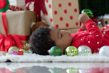 Portrait of cute little African kid boy in red clothes lying and playing with Christmas tree decorated balls on floor. Xmas tree and gift boxes in background. Xmas new year celebration concept