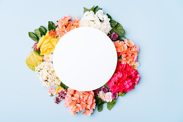 Blank paper card with round floral frame of mixed flowers on a pastel blue background. Top view of the model with copy space.