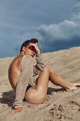 Beautiful fashion woman sitting on sandy beach, carefree moment. Stylish young female posing in knitted sweater relaxing on coast. Summer vacation