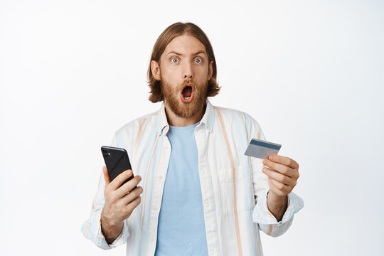 Image of surprised blond man gasp in awe, holding mobile phone and credit card, winning money, paying, making sale purchase, standing against white background