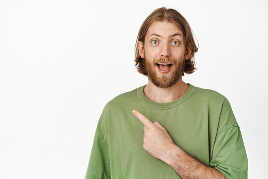 Excited bearded man gasp, wow face, pointing finger at upper left corner, intrigued by discount sale, has interest in advertisement, standing in green t-shirt against white background