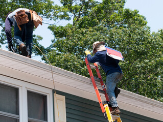 contractor climbing ladder with roof shingles while another nail the shingles on