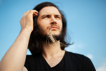 Outdoor portrait of young long haired man with wheat spike in the mouth.