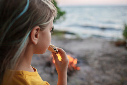 Cute girl roasting marshmallow to make smores over fire flame during camping, traditional American food, active recreation and family travel - Powered by Adobe