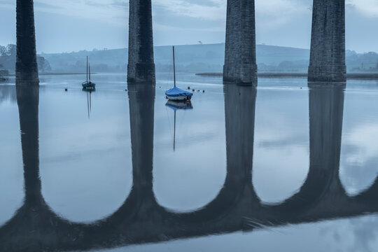 Boats moored beneath the towering arches of St. Germans viaduct over the River Tiddy at dawn in spring, St. Germans, Cornwall, England