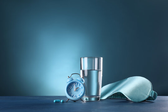 Alarm clock, soporific pills, sleeping mask and glass of water on blue wooden table, space for text. Insomnia treatment