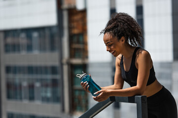 Side view of happy african american sportswoman looking at sports bottle on urban street