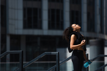 Side view of african american sportswoman with closed eyes holding sports bottle and jacket outdoors