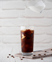 Iced coffee in a tall glass with cream on a white wood background. Cold tasty summer refreshment beverage concept. Selective focus, copyspace.