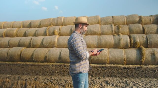 Farmer is standing beside bales of hay. He is examining straw after successful harvesting. Slow motion video.