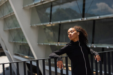 African american sportswoman in earphone standing with closed eyes near bridge on urban street