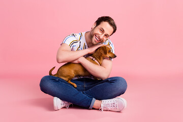 Full body photo of cheerful young happy man hold hands dog hug smile good mood isolated on pink...