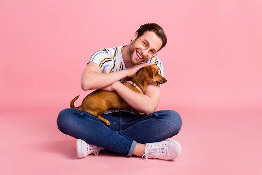 Full body photo of cheerful young happy man hold hands dog hug smile good mood isolated on pink color background