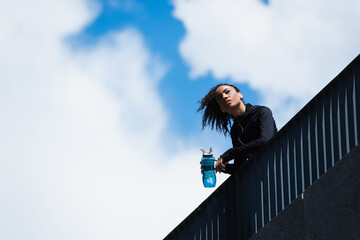 Low angle view of african american sportswoman holding sports bottle with sky at background