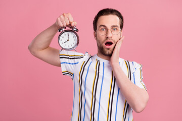 Photo of young amazed shocked man hold hand cheek alarm clock isolated on pastel pink color background