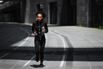 African american runner with sports bottle listening music in earphones on urban street