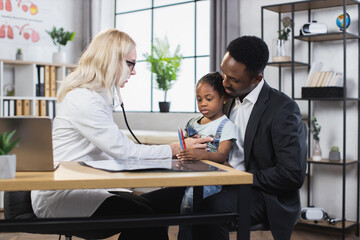 Caucasian female pediatrician listening heartbeat of little girl using stethoscope. African father with daughter visiting medical specialist for regular checkup.