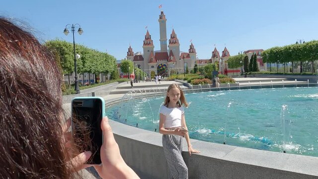 Mother holding smartphone and photographing daughter near fountain. Cropped shot of woman using mobile phone and taking photos of cute teenage girl near scenic outdoor fountain