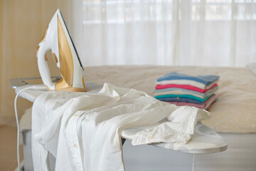 Neat stack of clean freshly laundered clothes and electric iron on pressboard in bedroom. Pile of...