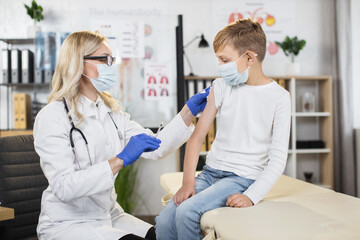 Obraz premium Medicine and vaccination concept during pandemic. Beautiful qualified blond woman doctor ready to prick needle into her patient boy's arm. Kid vaccination in medical center