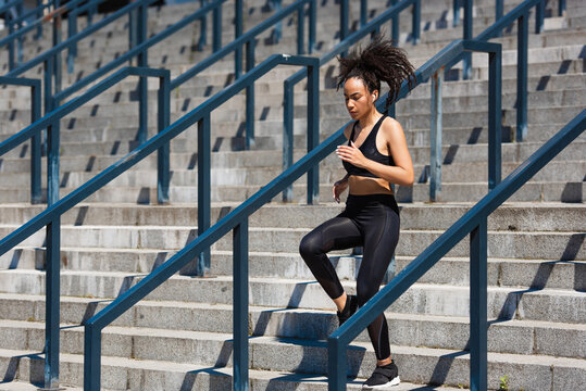 African american sportswoman in earphone running on stairs near railing on urban street