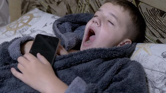Yawning Child in Home Gray Dressing Gown Falls asleep with Smartphone in Hands. A happy boy holds a mobile phone in his hands. A teenager is dependent on modern gadgets and tablets. 4K. Slow motion.