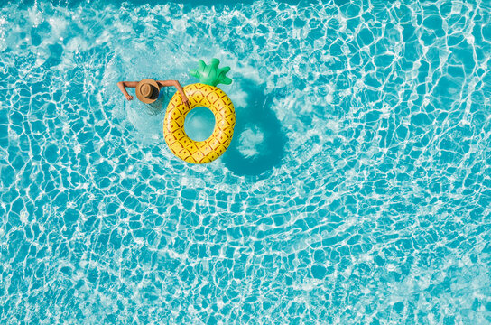Top view of a young female in swimsuit bikini in a straw hat on blue swimming pool waves background with big inflatable Yellow Pineapple tube. Chill out a summer vacation in luxury resorts concept.