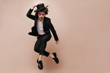Full-length portrait of pretty young gentleman in stylish shirt, black striped suit, classic hat and shoes, jumping while posing, crossing legs. Beige background