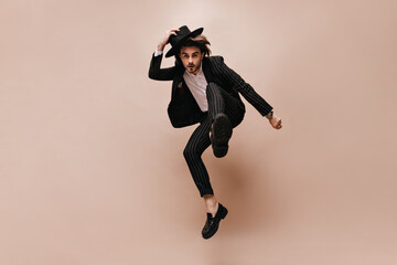 Fototapeta na wymiar Extraordinary posing in jump of young stylish man with brunette hair, white shirt, dark striped suit, black hat. Beige background in studio