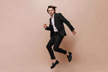 Fototapeta na wymiar Full-length photo of jumping young man with brunette hair, glasses, white shirt and black suit. Model looking into camera and posing against beige background