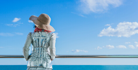 a woman with her back turned and wearing a hat looks at the sea on a blue sky day.