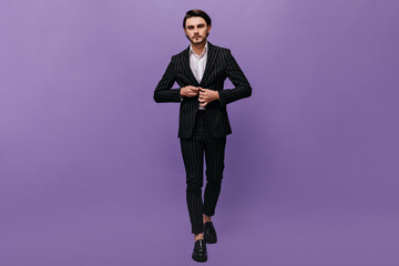 Obraz na płótnie Canvas Full-length photo of sexy young man with brunette hair, classic striped suit, white shirt and black shoes, fastening a button on jacket. Model looking into camera against violet background
