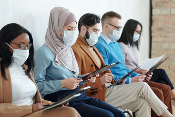 Fototapeta na wymiar Multiracial people in protective masks sitting in corridor and looking through their resume
