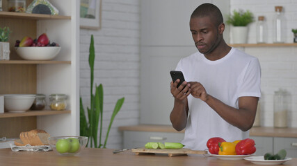 African Man using Smartphone while Cutting Vegetables in Kitchen