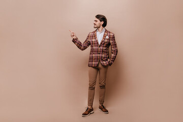 Full-length portrait of elegant young man in white shirt, plaid retro blazer and pants, turning head, pointing and looking aside at copy space, posing isolated over beige background