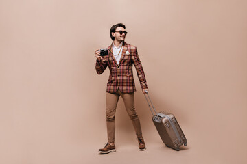Stylish voyager in beige pants, brown leather shoes, plaid jacket, holding camera and suitcase,...