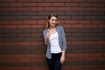 Half length portrait of carefree charming woman dressed in casual clothing looking at camera and showing thumbs up, attractive female student posing near brick wall with advertising copy space area