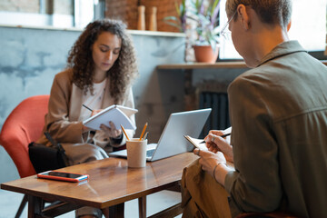 Two businesswomen making notes in notebooks by table in office