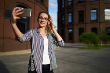 Millennial hipster girl in optical spectacles for provide eyes correction using cellphone front camera for clicking images connecting to 4g internet during vacations for travelling, selfie influence
