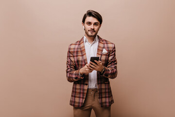 Stunning young gentleman with brunette hair and bristle, wearing white shirt, plaid blazer and brown pants, looking into camera and holding phone. Man posing over beige background