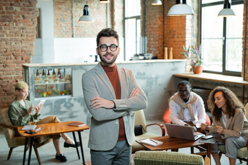 Young smiling cross-armed businessman standing in front of working colleagues