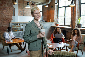 Pretty businesswoman with drink looking at camera in office where her colleagues using mobile gadgets