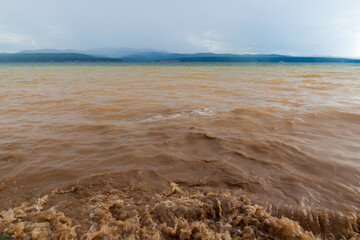 Muddy brown water with sand on the surface of the lake. Cloudy weather after storm