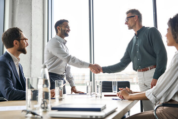 Naklejka na ściany i meble Two happy diverse professional business men executive leaders shaking hands after successful financial deal at group board office meeting. Trust agreement company trade partnership handshake concept.