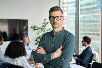 Smiling confident mature businessman leader looking at camera standing in office at team meeting. Male corporate leader ceo executive manager wearing glasses posing for business portrait arms folded. - obrazy, fototapety, plakaty