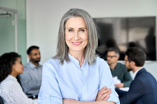 Happy Older Senior Woman Business Leader Standing In Office Working With Team People. Smiling Middle Aged 50s Professional Executive, Mentor Or Manager Posing For Portrait With Arms Crossed.