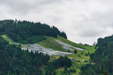 Solar park, photovoltaic power station located on a mountain slope in the Alps. Foggy day, green pasture grass and blue skies and mountains create a fresh and ecological image. - obrazy, fototapety, plakaty