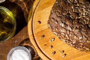 Still life - black bread with sunflower seeds, olive oil and coarse salt in glass jars, a knife, and a linen napkin on a wooden board, wooden background, hard light, photo in a low key.