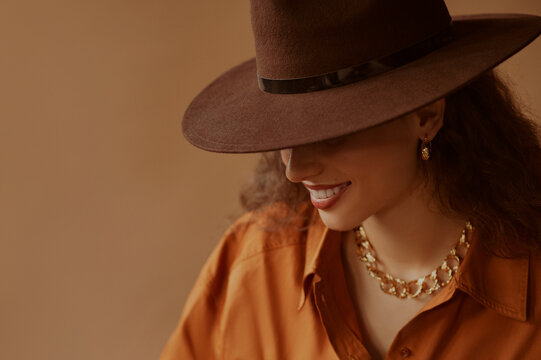 Beautiful happy smiling woman  hiding her face under trendy brown hat, wearing chunky golden chain, stylish shirt, posing on beige background. Autumn fashion conception. Copy, empty space for text
